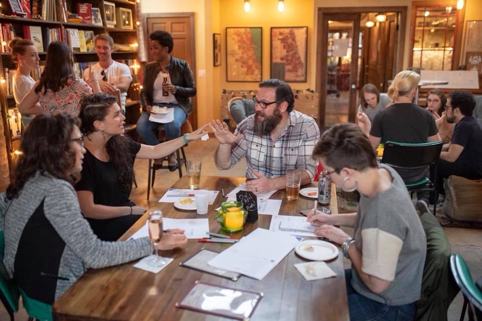 An image of a ballot party with some participants engaged in conversation around a table and others taking notes on paper